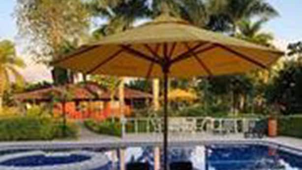 An outdoor pool area with a table and chairs under a large umbrella, surrounded by palm trees and a building in the background. - Villa Thiago Finca Tradicional (Armenia)