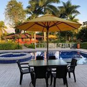 An outdoor pool area with a table and chairs under a large umbrella, surrounded by palm trees and a building in the background. - Villa Thiago Finca Tradicional (Armenia)