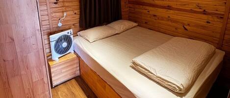 A wooden cabin bedroom with a bed, a nightstand, a fan, and a window with curtains.
