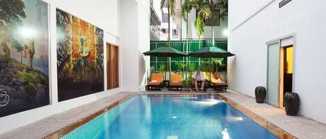 A swimming pool with two large vases at the end, surrounded by lounge chairs and umbrellas, with artwork on the walls and palm trees in the background.