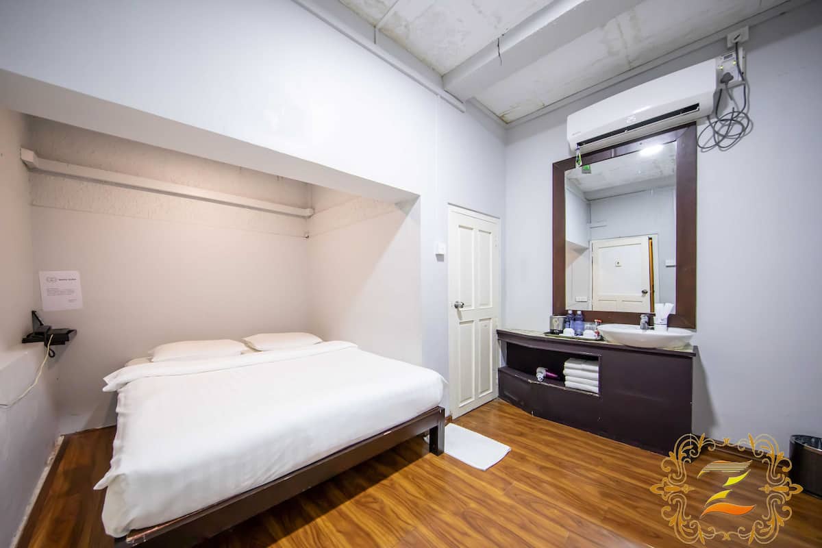 Una camera da letto con un letto singolo, pavimento in legno, parete bianca, porta bianca, uno specchio e un piccolo ripiano con articoli da toilette.