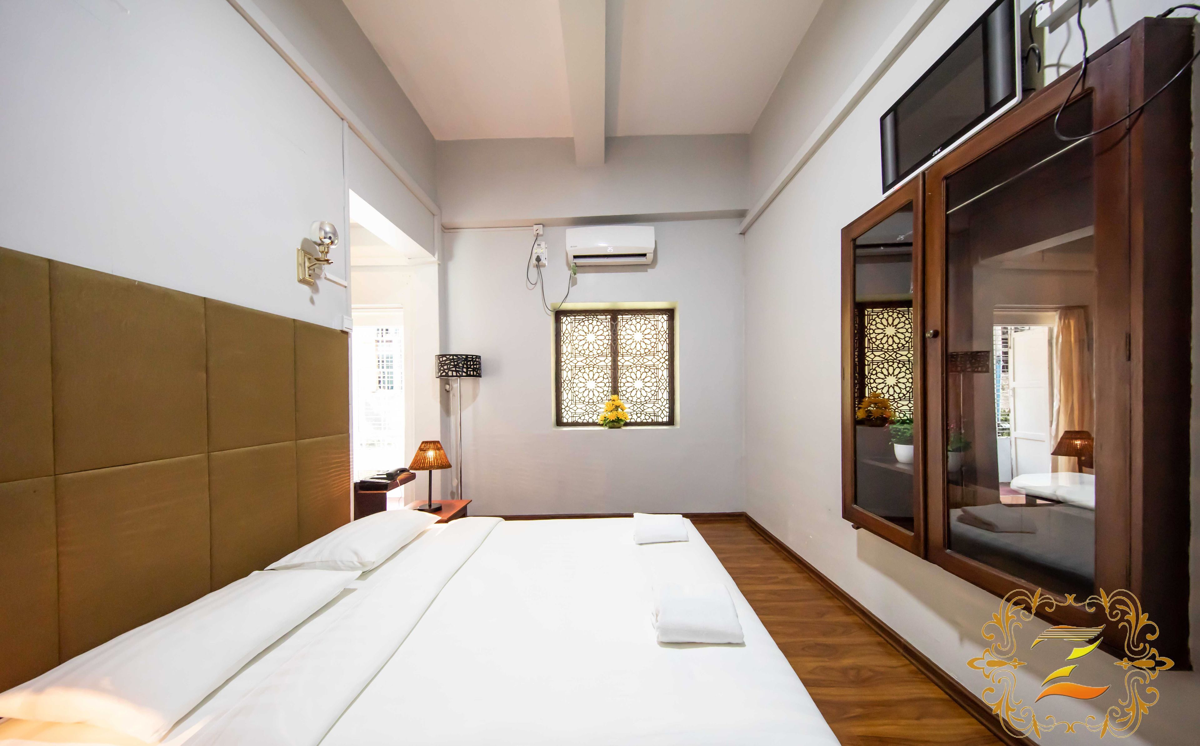 A hotel room with a bed, a nightstand, a lamp, a window with decorative screen, an air conditioning unit, and a television on a wooden stand.