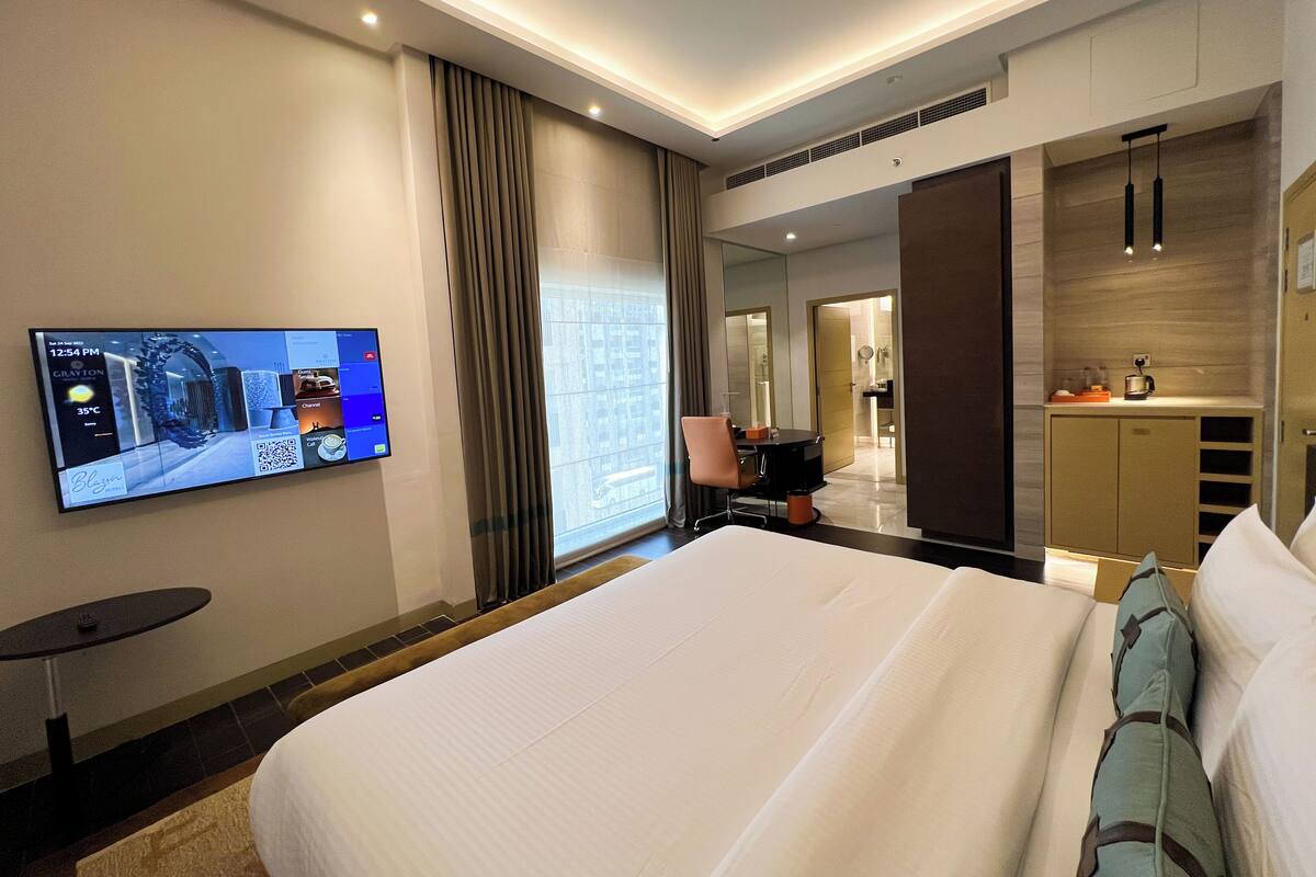 A hotel room with a large bed, a desk with a chair, a TV mounted on the wall, and a view of the city through the window.