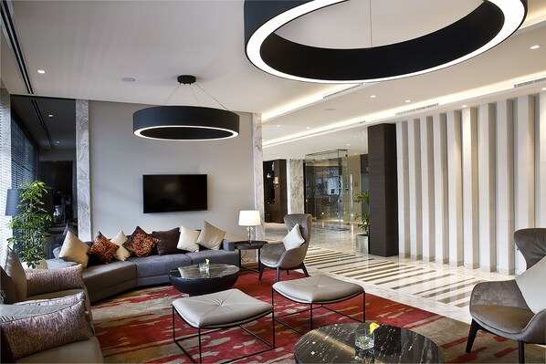 A modern hotel lobby with a curved ceiling light, a flat-screen TV, a sofa, armchairs, and a coffee table.