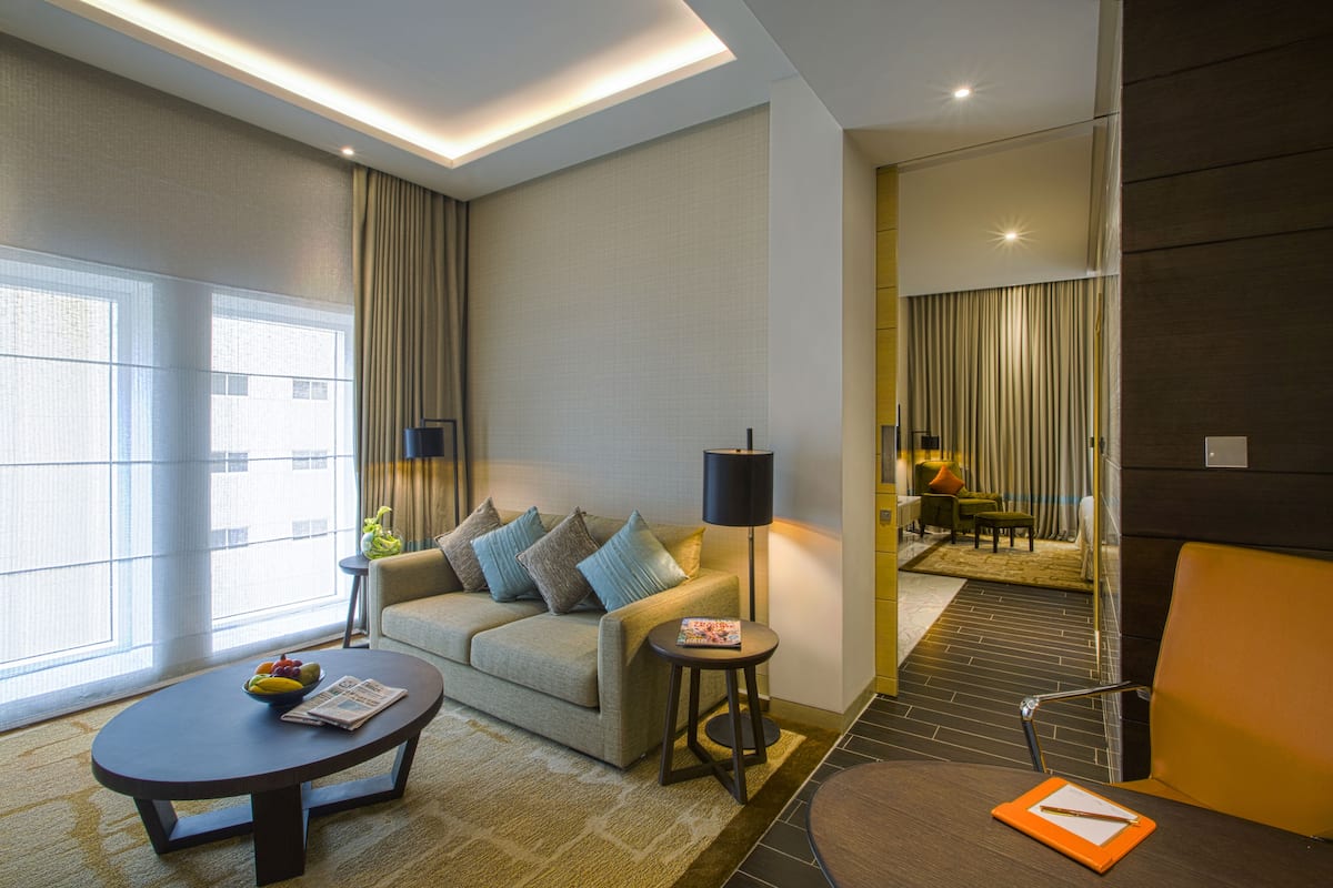 A modern hotel room with a sofa, a small round table, a lamp, and a view of the city through large windows.