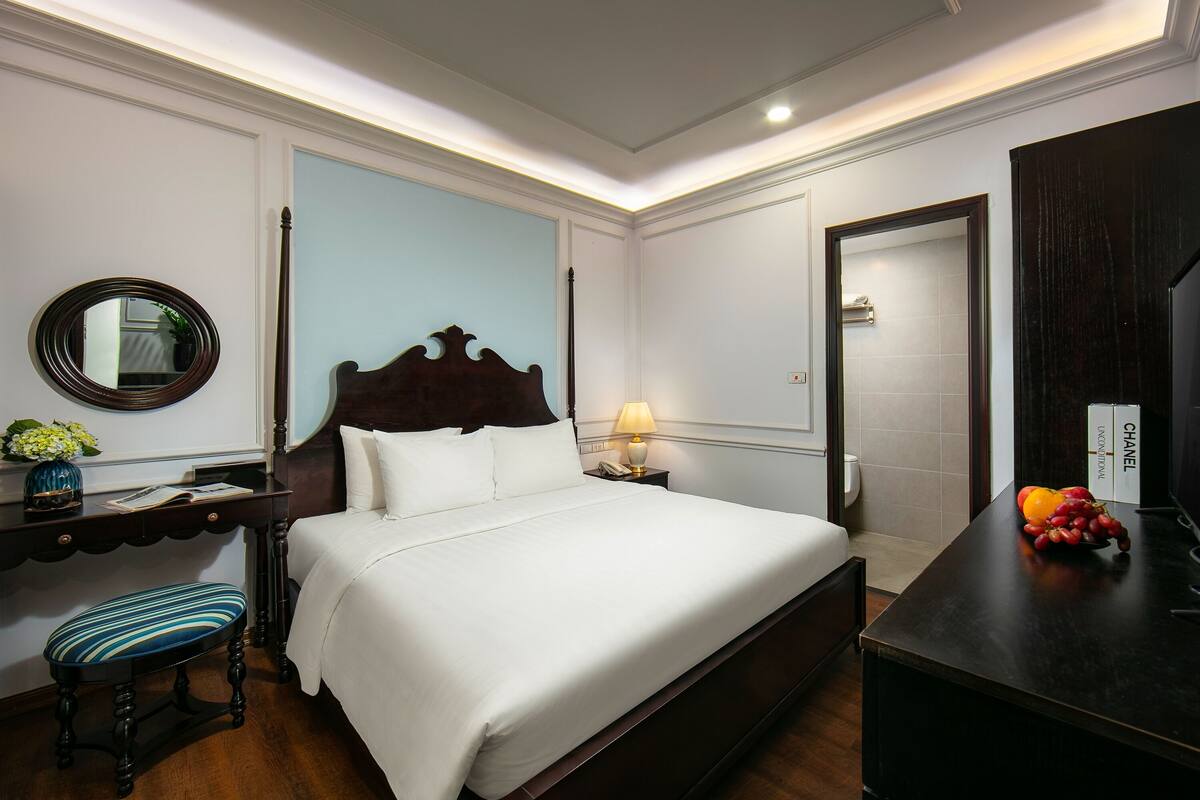 Une chambre d’hôtel avec un grand lit, une table de chevet avec une lampe, un petit tabouret, un miroir et un panier de fruits.