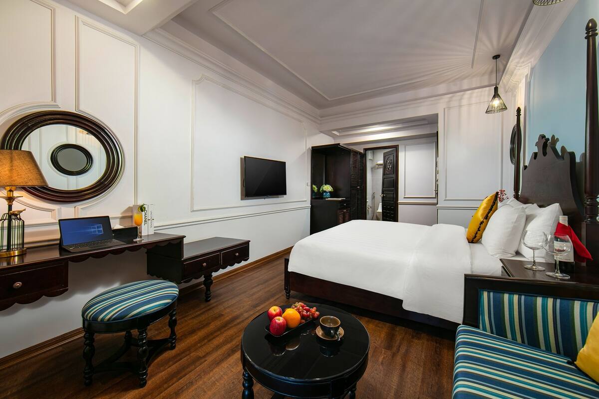 Une chambre d’hôtel avec un lit, un bureau avec un ordinateur portable, un miroir rond, une petite table ronde avec des fruits et une tasse, et un canapé.