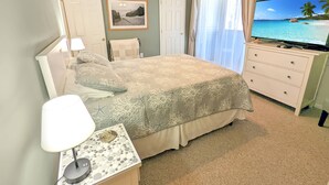 Une chambre à coucher comprenant un lit, un téléviseur, un ventilateur de plafond, un bureau avec une lampe et une fenêtre avec des rideaux.