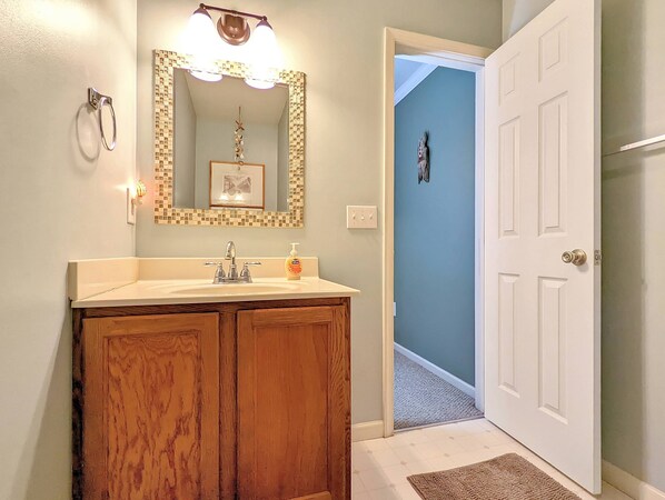 Une salle de bain avec un meuble-lavabo en bois, un lavabo blanc, un miroir avec un cadre en mosaïque et une porte donnant sur une autre pièce.