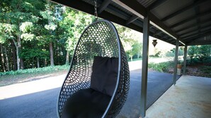 Een hangstoel met een zwart kussen onder een overdekt terras met een betonnen vloer en metalen balken.