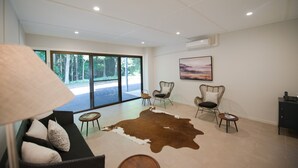 Ruime kamer met groot raam, een kuitenvacht vloerkleed, rieten stoelen en een schilderij aan de wand.