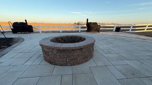 Un foyer en pierre est installé au centre d’une terrasse dallée, avec une clôture blanche en arrière-plan.