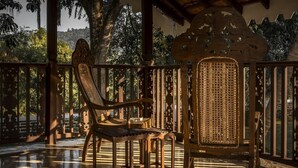 Una silla de madera con asiento y respaldo trenzados, ubicada sobre un suelo de baldosas, con vista a árboles y un paisaje montañoso a través de una baranda de madera.