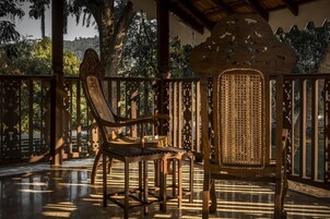 Una silla de madera con asiento y respaldo trenzados, ubicada sobre un suelo de baldosas, con vista a árboles y un paisaje montañoso a través de una baranda de madera.