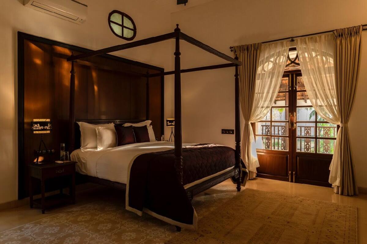 A four-poster bed with a dark wooden frame, a large window with curtains, and a nightstand with a lamp.