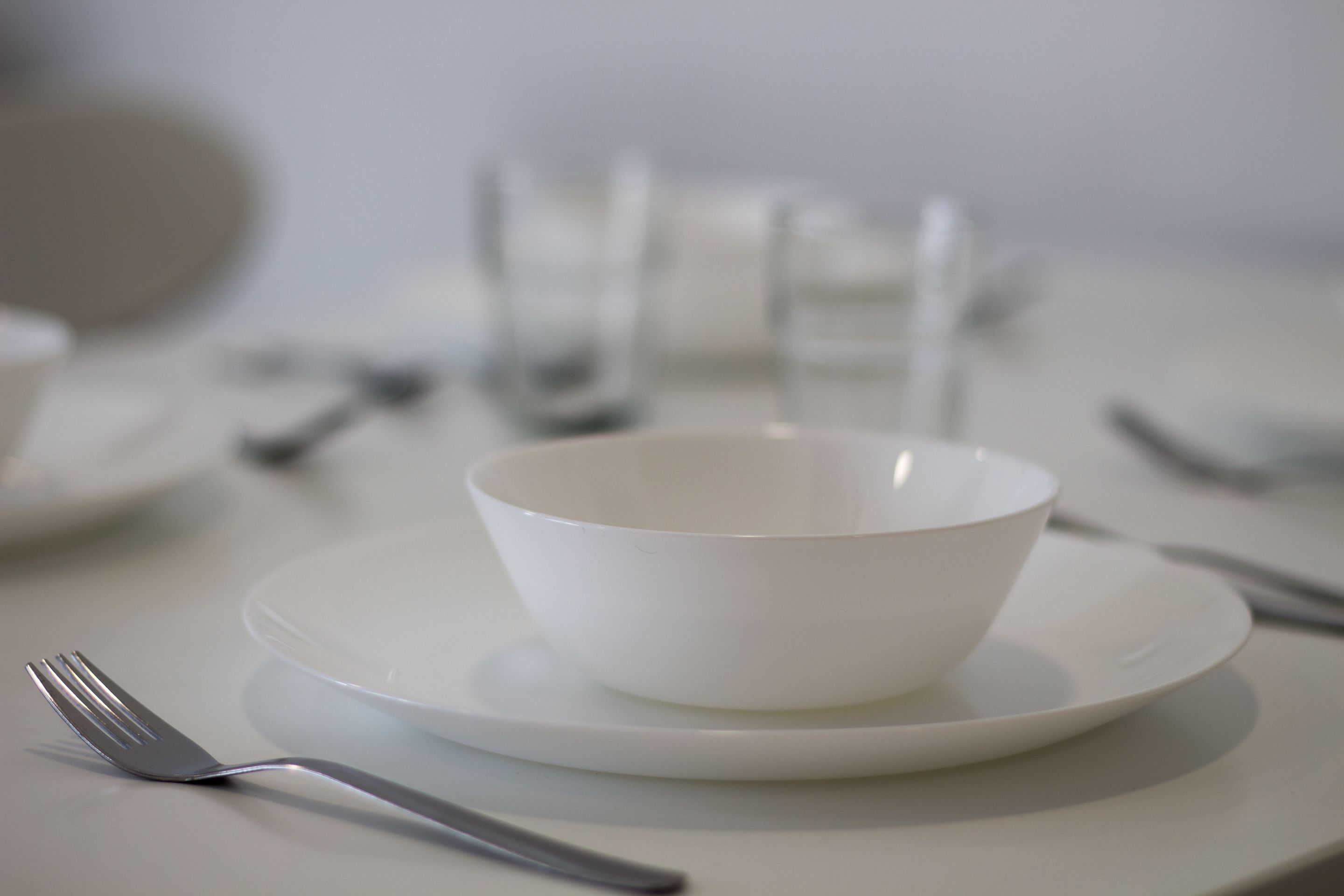 Una mesa de comedor blanca con un bol y un plato blancos, un tenedor y una copa.