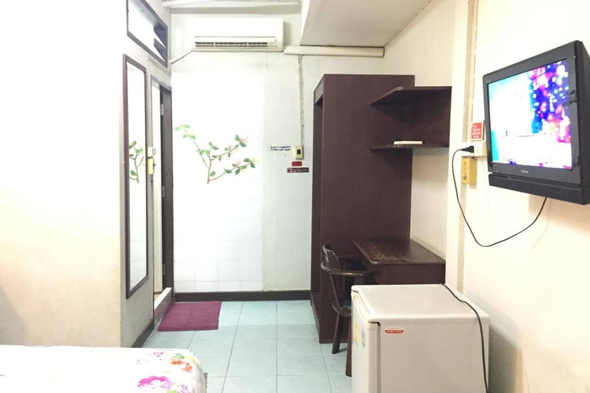 A narrow hallway with a bed, a small refrigerator, a TV mounted on the wall, and a door leading to another room.