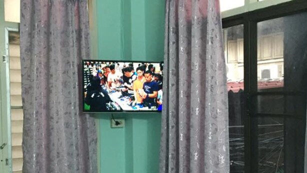 Ein Zimmer mit türkisfarbenen Wänden, einem Bett mit blumigem Bettbezug, einem an der Wand montierten Fernseher und einem Deckenventilator.