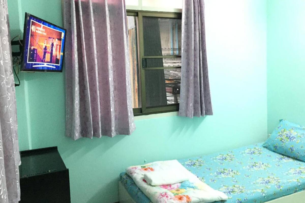 A small room with a single bed, a window with curtains, a television on the wall, and a black cabinet.