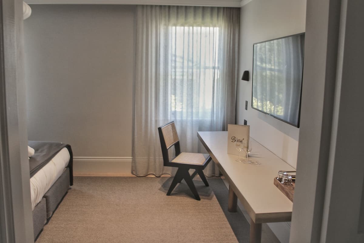 A bedroom with a bed, a desk, a chair, a window with curtains, and a mirror on the wall.
