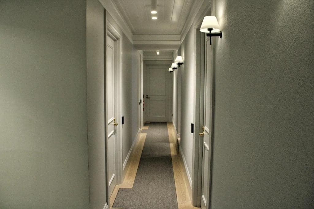 Un couloir avec des portes de part et d’autre, un sol recouvert de moquette et des luminaires fixés aux murs.