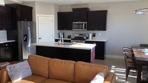 A modern kitchen with dark cabinets, a stainless steel refrigerator, a microwave, and a gas stove. There is a dining area with a table and chairs, and a brown leather sofa with a fluffy throw. - MONTHLY RENTAL ONLY  (Hurricane)