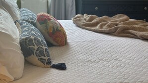 A neatly made bed with a quilted bedspread and several decorative pillows. A beige throw blanket is draped over the bed. In the background, there is a dark wooden dresser with a mirror above it, and a window with curtains allowing natural light into the room. - Charming Condo in Downtown Wilmington’s Historic District (Wilmington)