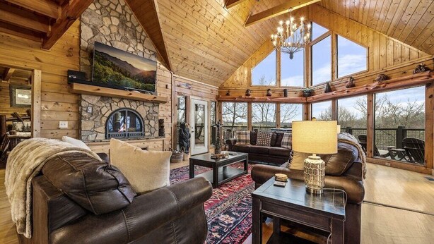 A cozy log cabin interior with a stone fireplace, leather furniture, and large windows offering views of the outdoors.