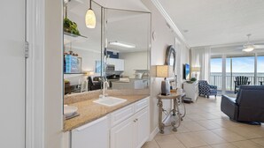 A modern kitchen with a large island, a dining area with a table and chairs, a living room with a sofa, and a view of the ocean through large windows. - Regency Isle 608 by Liquid Life (Orange Beach)