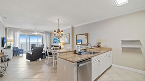 A modern kitchen with a dishwasher, a dining area with a table and chairs, and a living room with a sofa and a TV. - Regency Isle 608 by Liquid Life (Orange Beach)