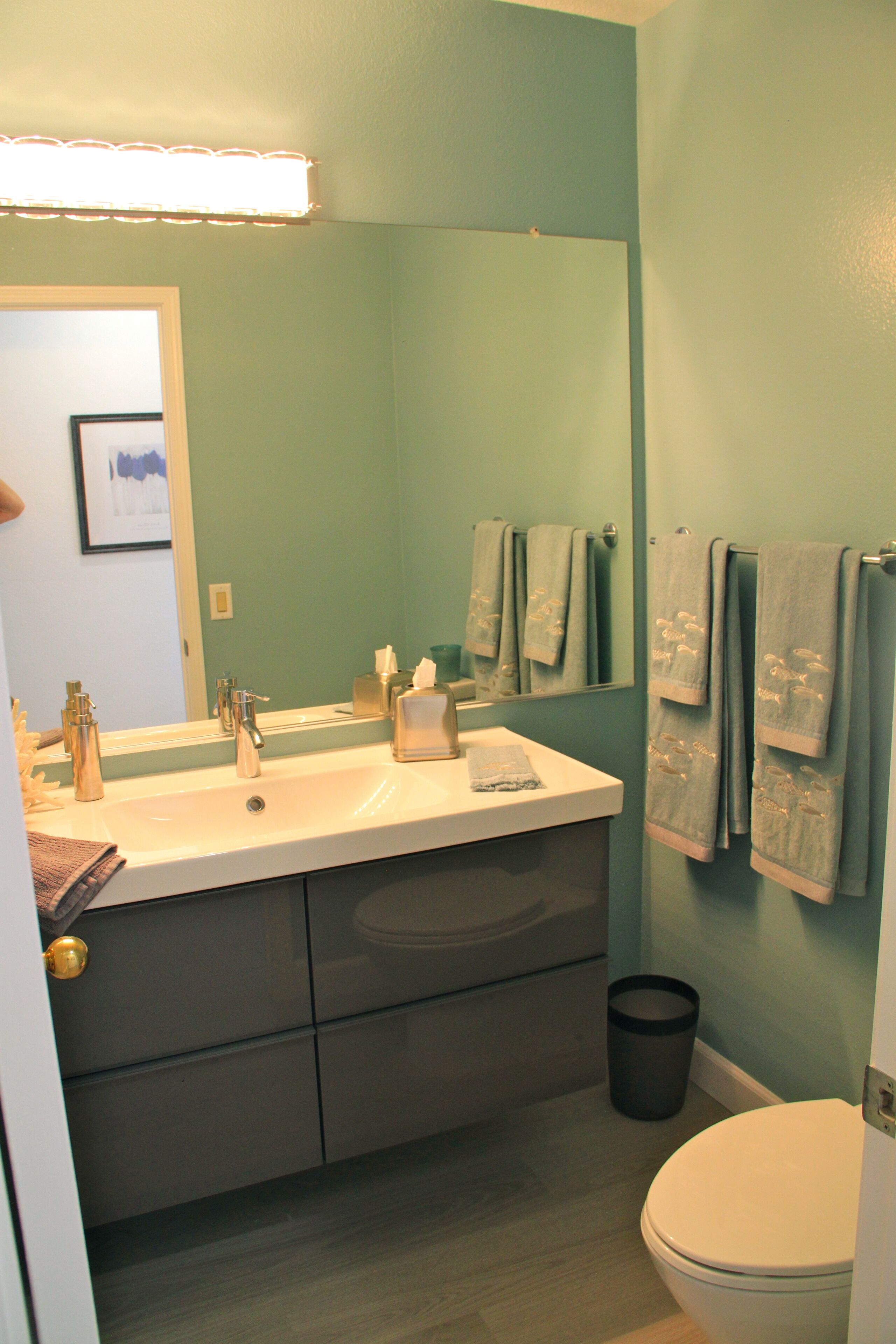 A bathroom with a double sink, a mirror, towels, a trash can, and a toilet.