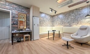Habitación de hotel con piso de madera, pared de mármol, un sillón blanco, una mesita pequeña y un escritorio con espejo.