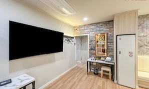 Habitación de hotel con televisor de pantalla plana en la pared, una pequeña mesa con una silla, minibar y una puerta con un cartel que indica el nombre y el año del hotel.