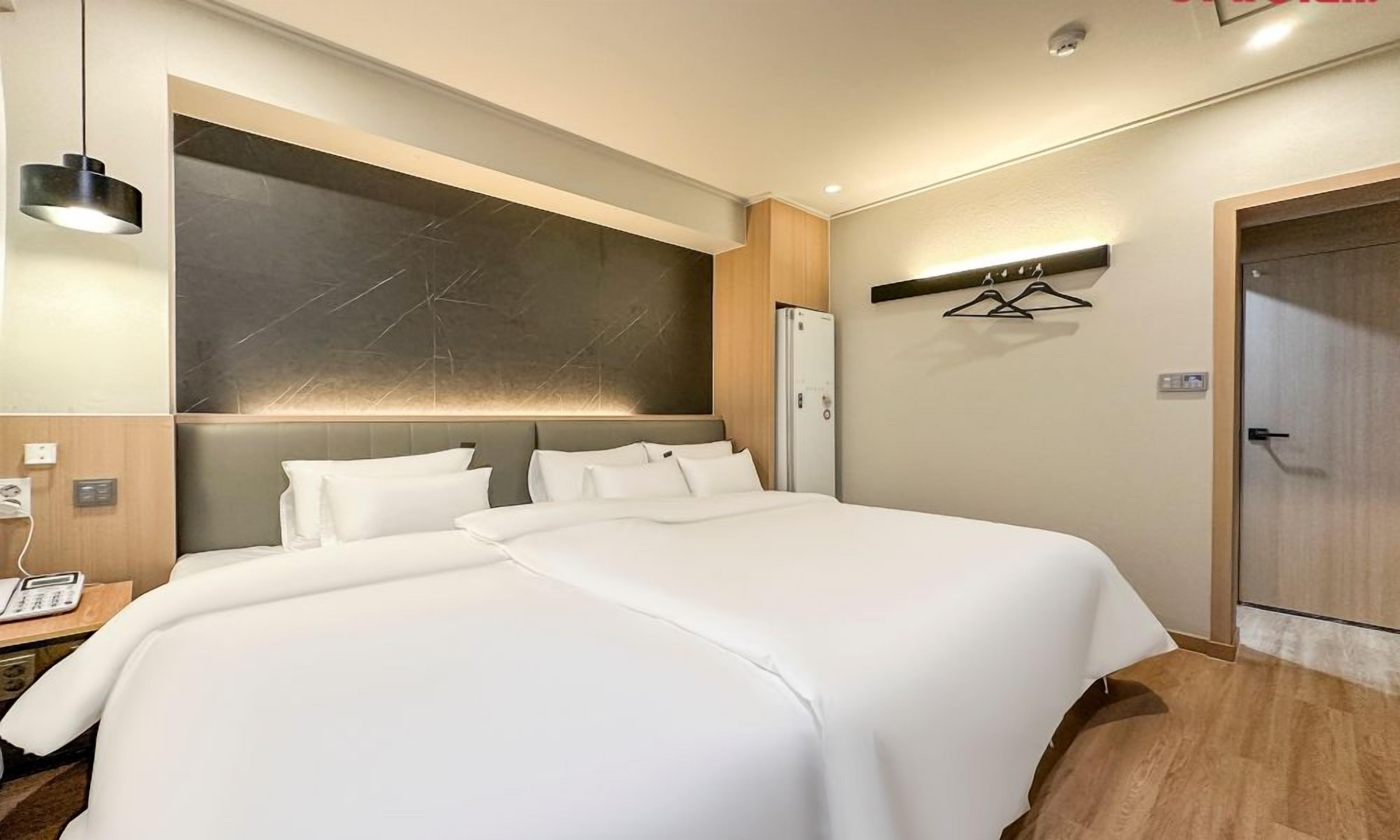 A hotel room with a large bed, a bedside table with a phone, a wall-mounted light, and a door with a coat rack.