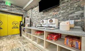 Zona de recepción de un hotel con una pared de ladrillo, un televisor de pantalla plana sobre el mostrador y estantes llenos de aperitivos.