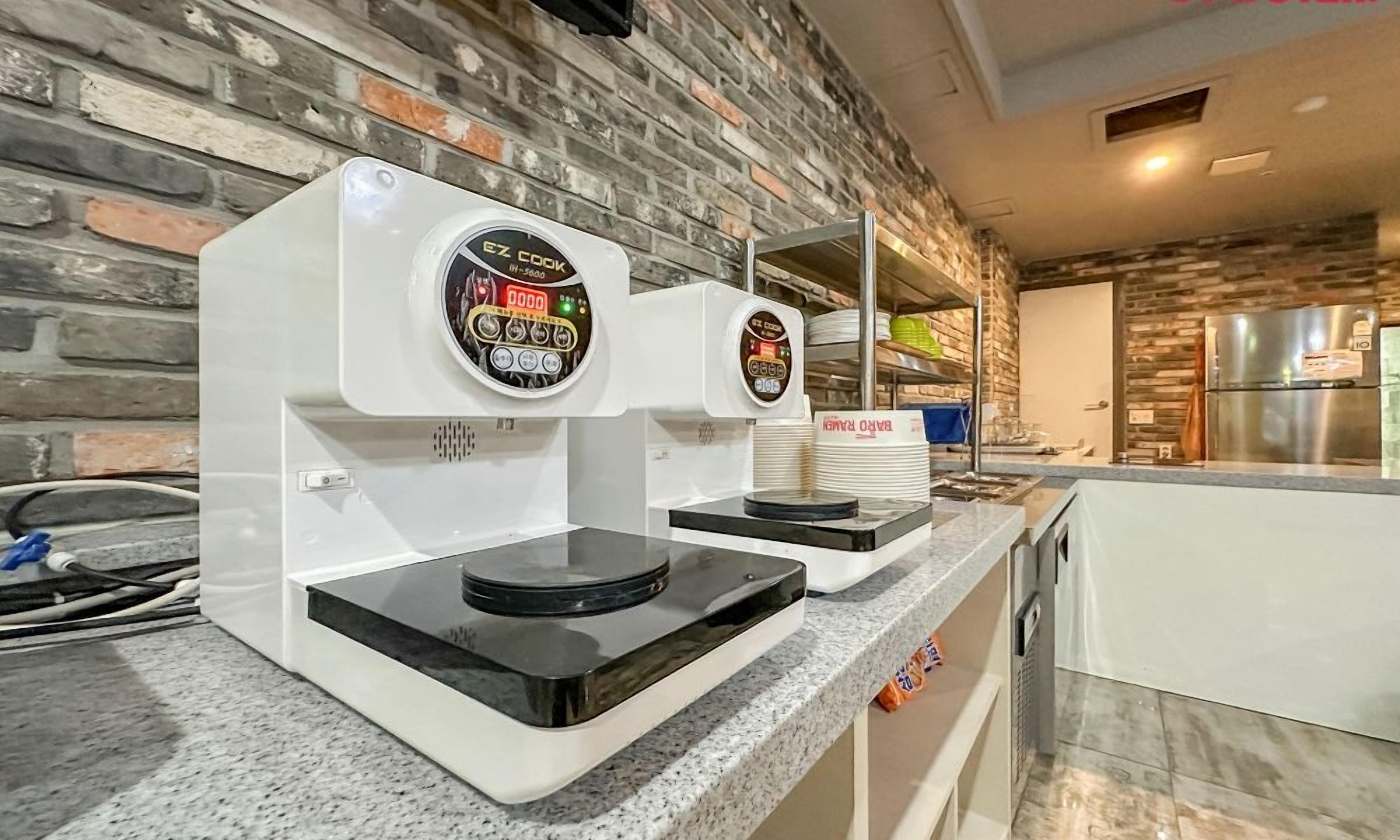 A kitchen with two coffee machines on a granite countertop, a brick wall, and a stainless steel refrigerator in the background.