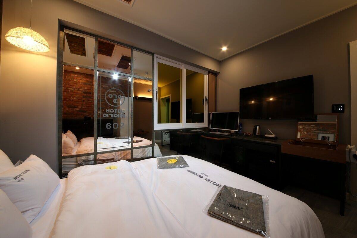 Habitación de hotel con cama, escritorio, televisor y puerta corredera de vidrio que da a un baño.