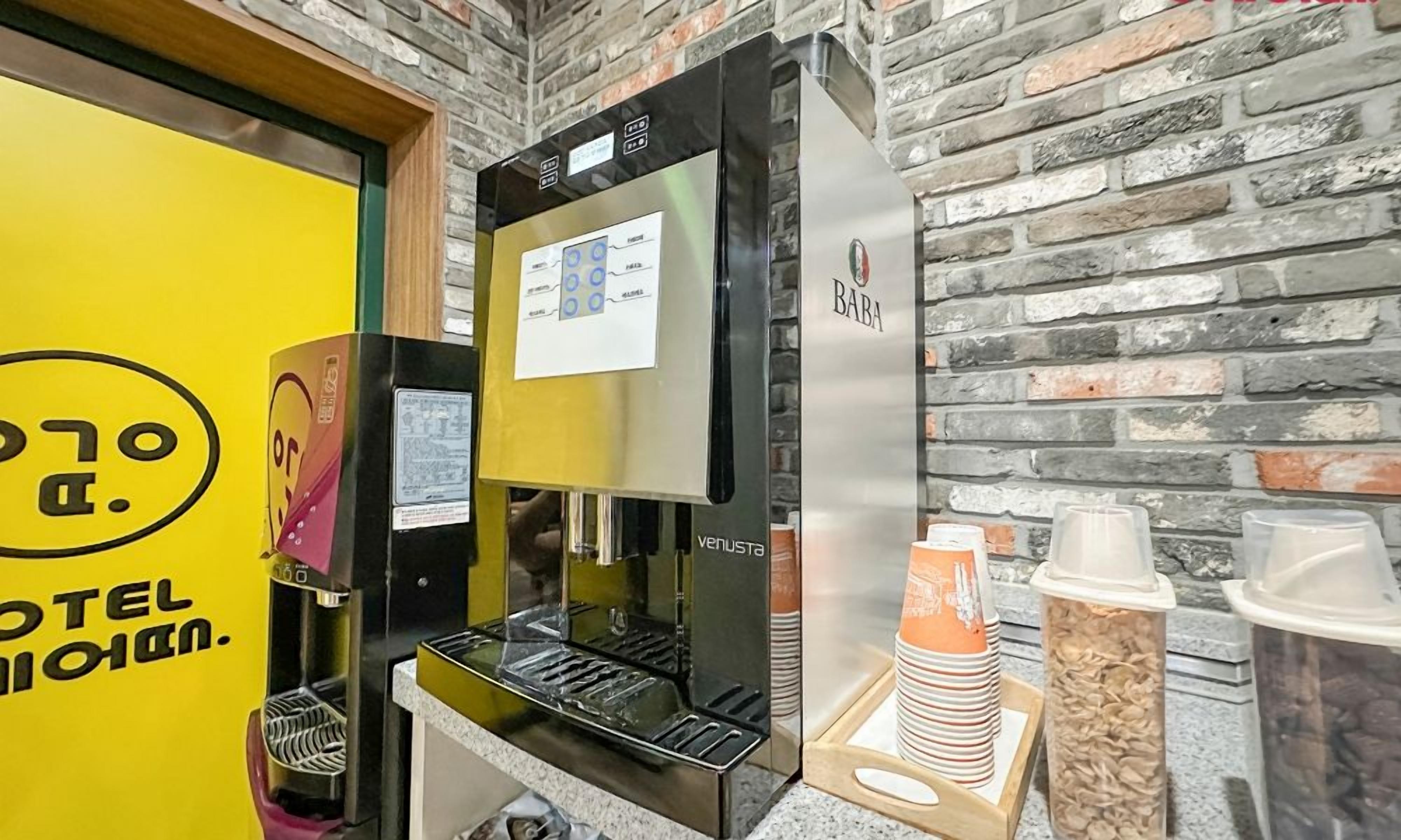 A coffee machine with a digital display, a yellow wall with a logo, and a brick wall background.
