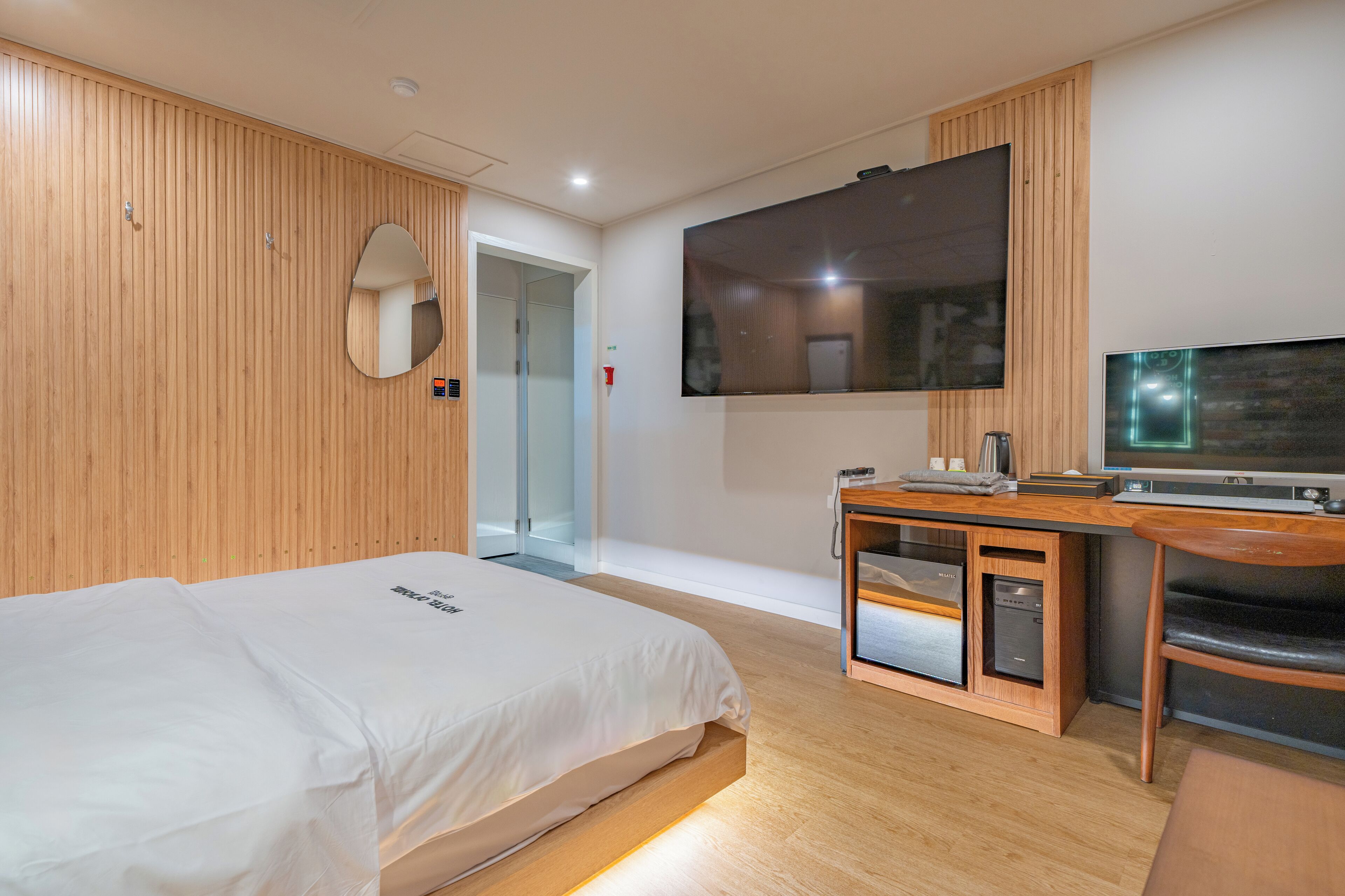 Habitación de hotel moderna con pared de paneles de madera, cama con colcha blanca, escritorio con televisor de pantalla plana y lavavajillas integrado.