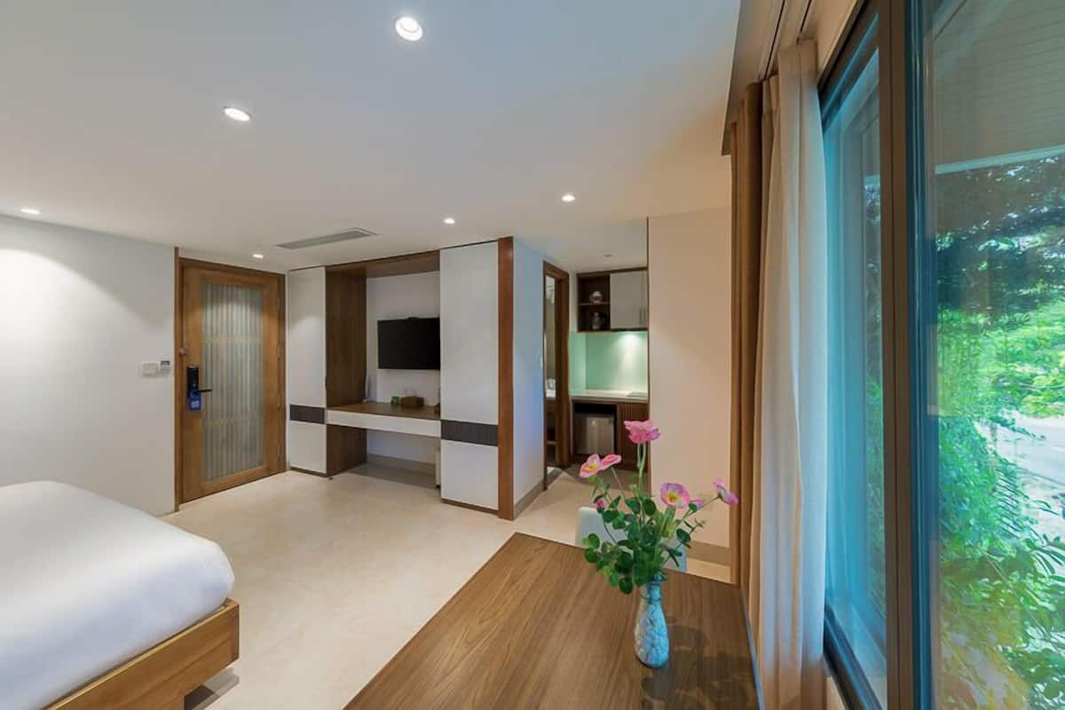 A modern hotel room with a bed, a desk, a TV, and a large window with a view of greenery.