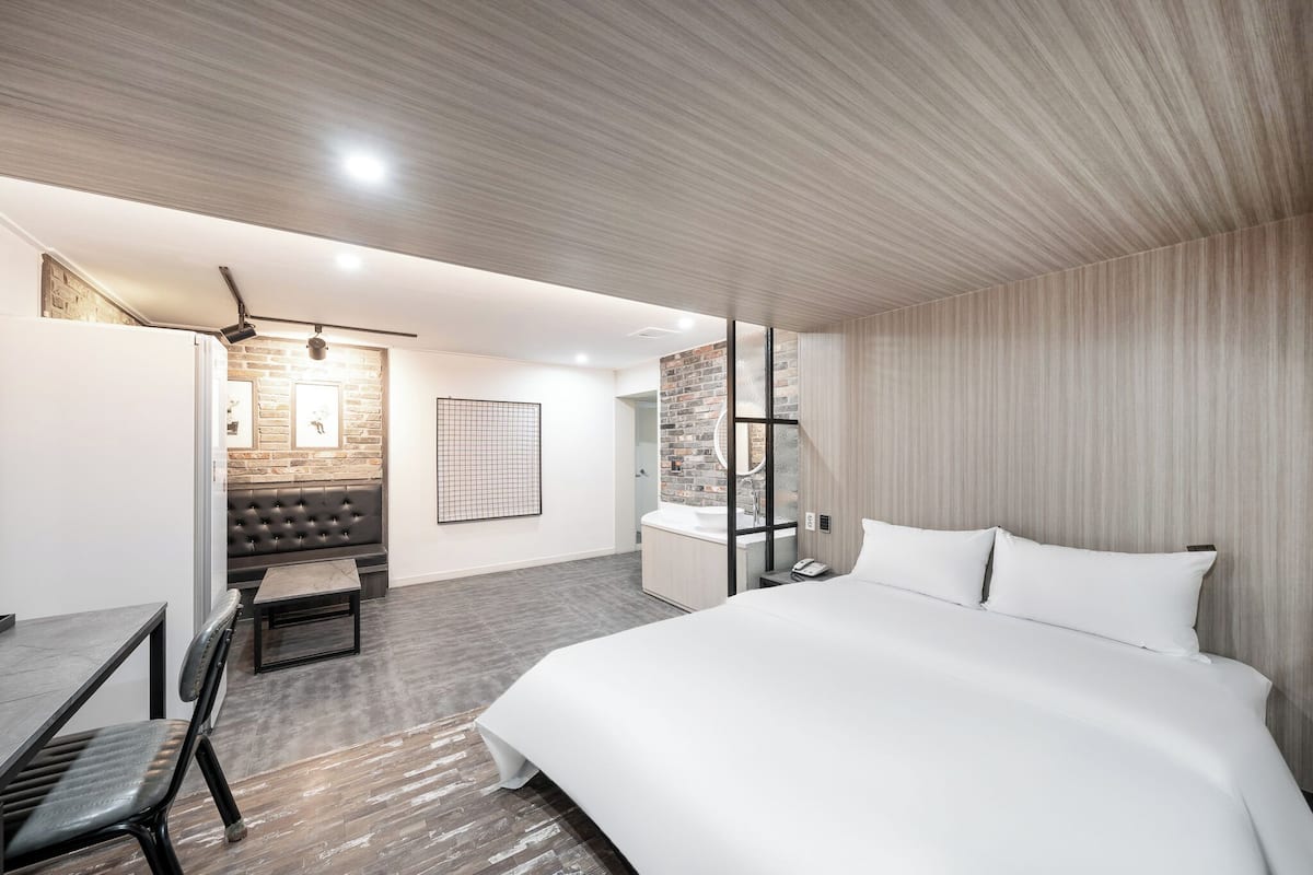 A modern hotel room with a large bed, a seating area with a sofa and table, a bathroom with a glass shower, and a brick accent wall.