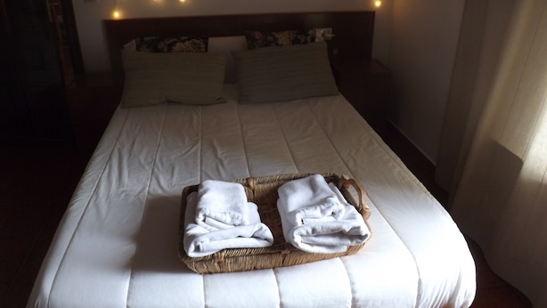 Un lit bien fait, avec du linge de lit blanc, une tête de lit ornée d’une guirlande lumineuse décorative et un panier en osier contenant des serviettes pliées, posé sur le lit.