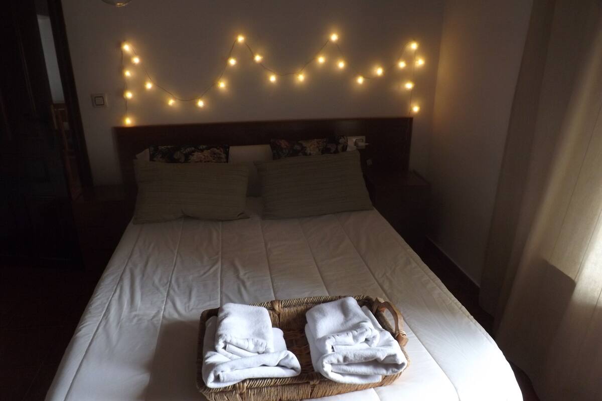 Ein ordentlich bezogenes Bett mit weißen Leinen, ein Kopfteil mit einer dekorativen Lichterkette und ein gewebter Korb mit gefalteten Handtüchern auf dem Bett.