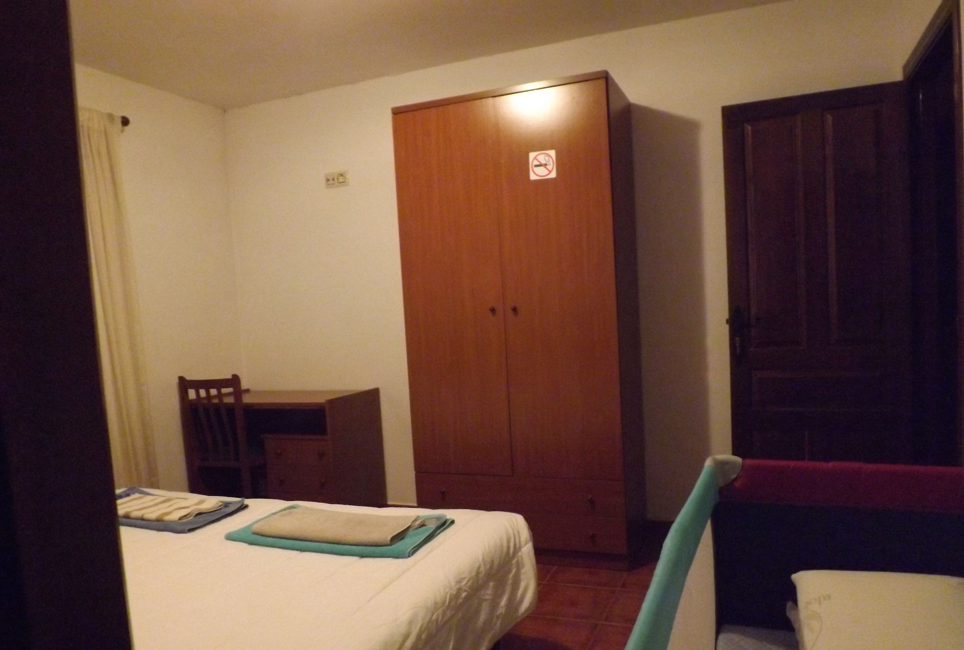 Una habitación pequeña y con poca luz, con una cama individual, un armario de madera y un escritorio con una silla.