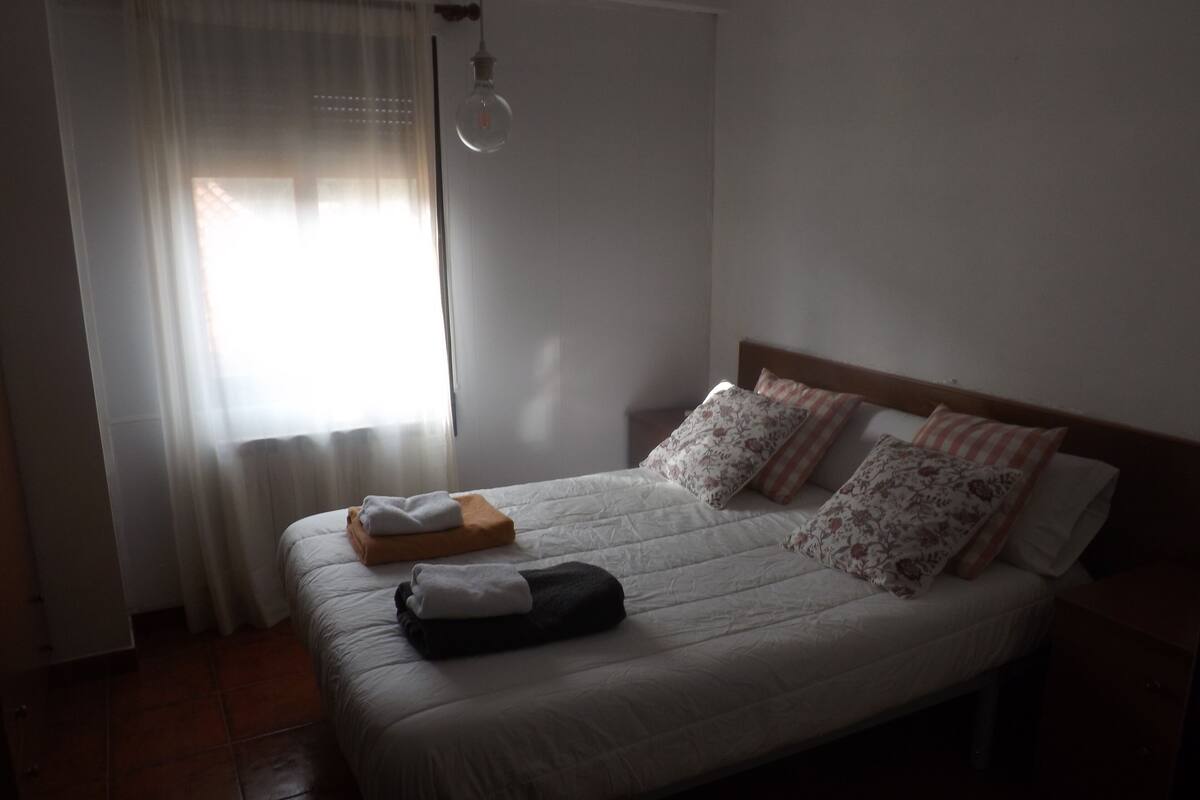 Ein ordentlich bezogenes Bett mit weißer Decke und mehreren Kissen, ein Holz-Kopfstück sowie ein Nachttisch mit Lampe.