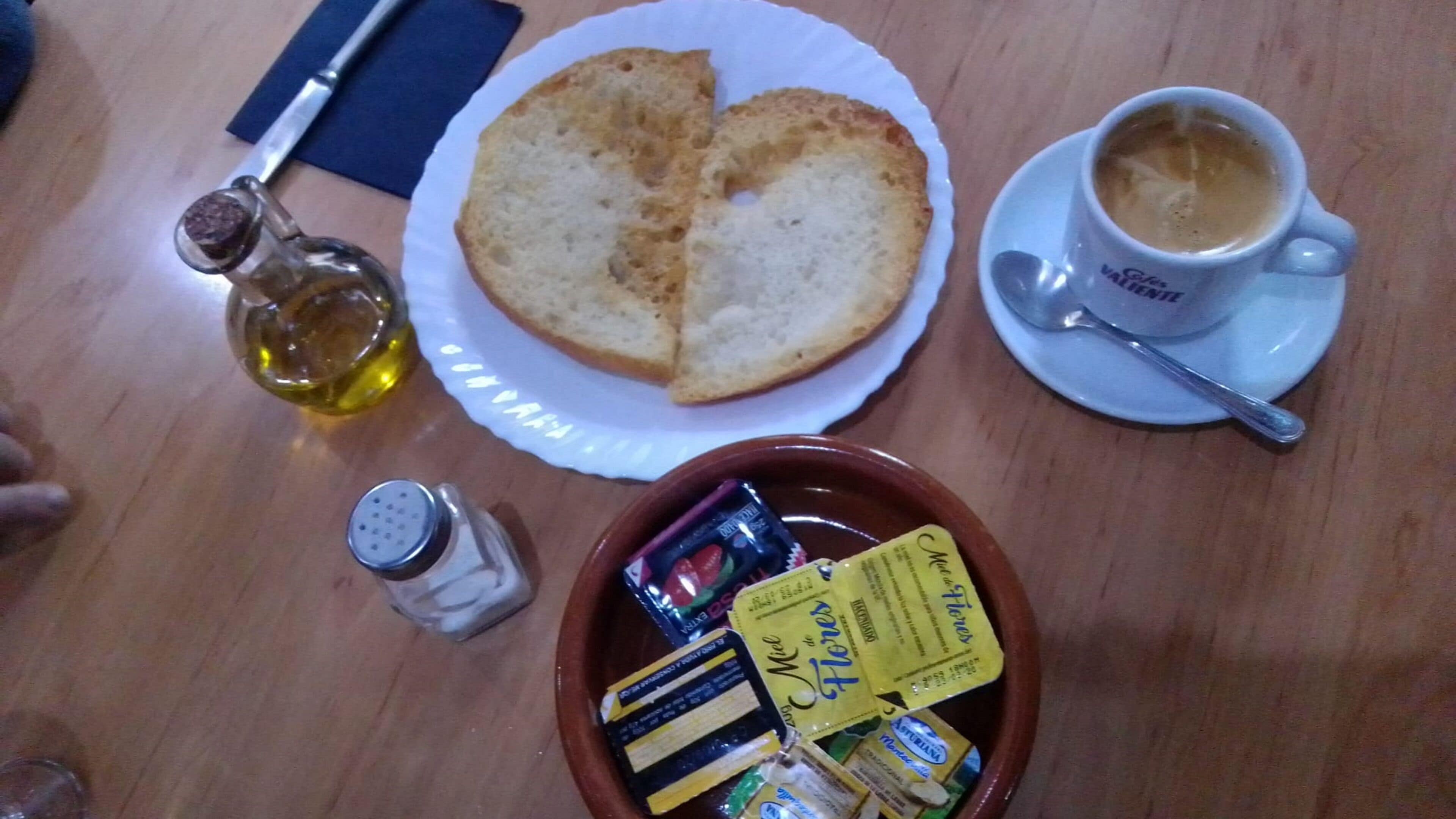 Un tavolo con una fetta di pane tostato, una tazza di caffè, una bottiglia d'olio, un saliera, una pepe e una ciotola di snack assortiti.