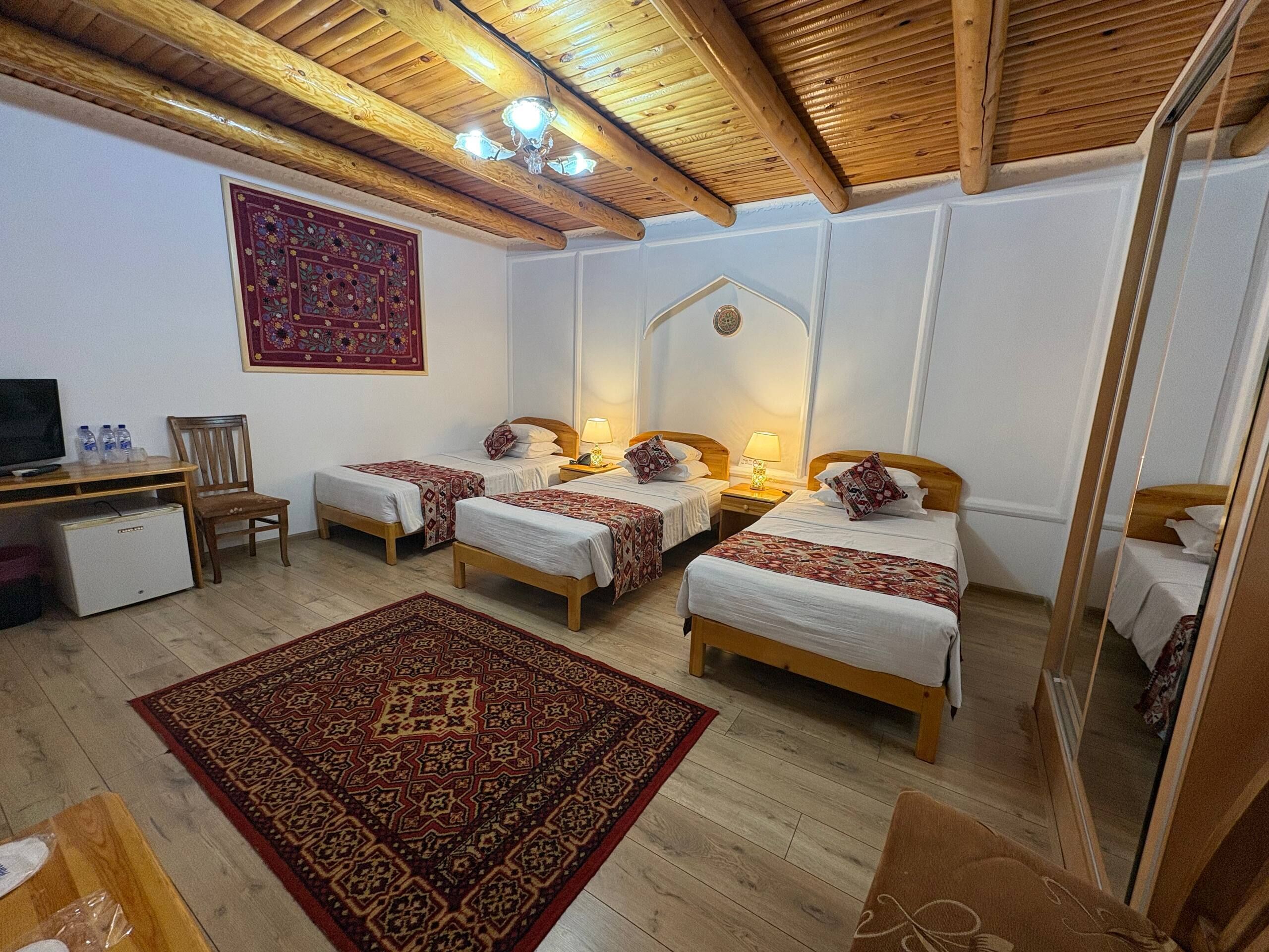 Une chambre avec quatre lits simples, un bureau avec une télévision, un réfrigérateur et un grand tapis à motifs.