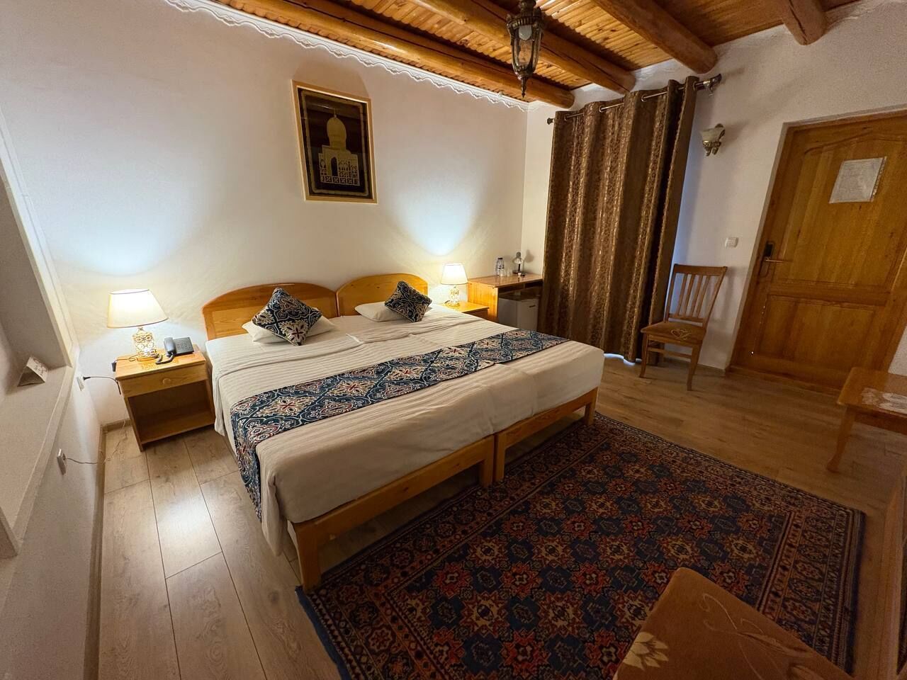 A bedroom with a double bed, wooden headboard, nightstands, a bedside lamp, a patterned rug, a wooden door, and a chair.
