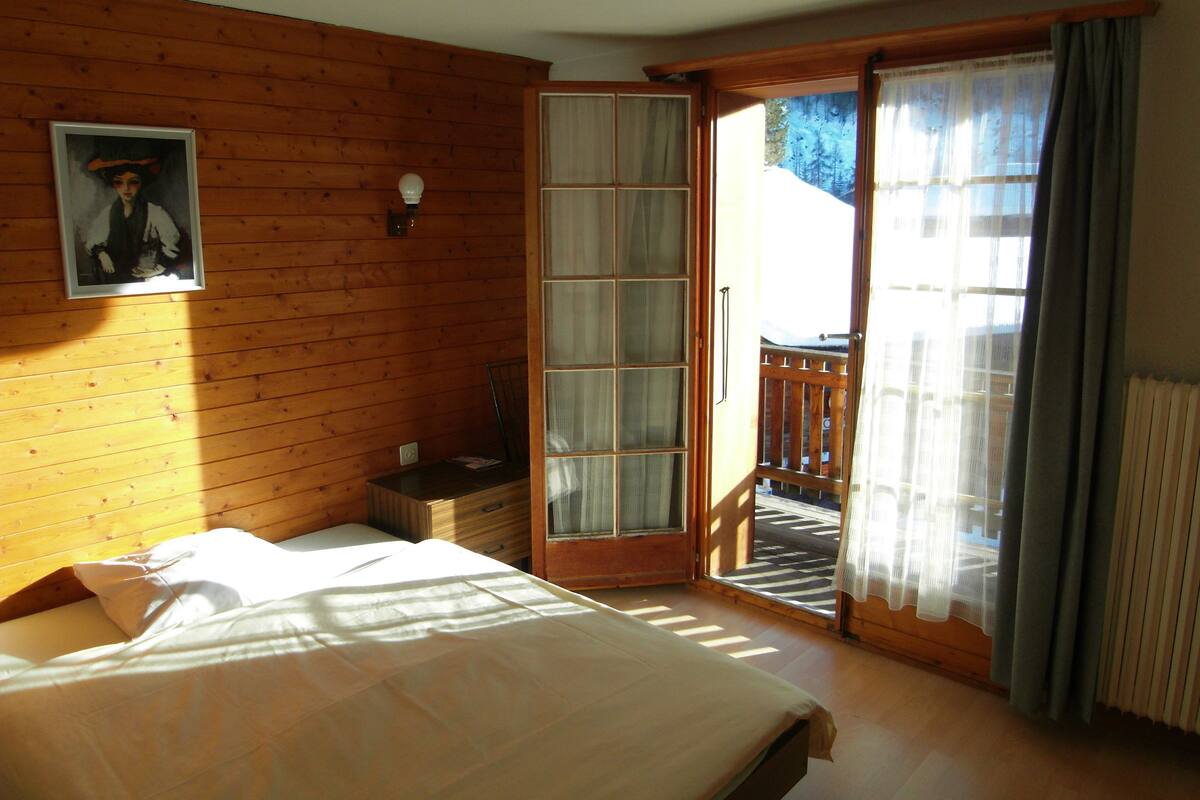 A bedroom with a bed, a nightstand, a window with a view, and a door leading to an outdoor area.