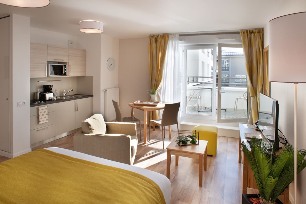 A modern hotel room with a bed, a desk, a chair, a small table, a microwave, a sink, and a balcony with curtains.